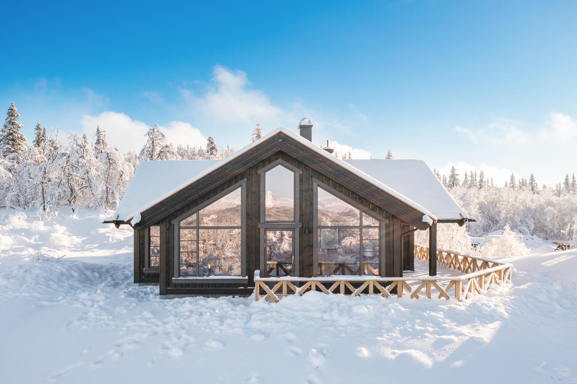 A newly built cabin with dark walls and large windows, deep in fresh snow under a blue sky